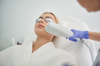 Close up of girl in white bathrobe lying on daybed while beautician in sterile blue gloves using laser epilation machine to remove unwanted hair, wrinkles or skin irregularities on her face