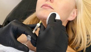 Woman gets volume to chin, reshapes with filler, injection. White female lying on couch. Beauty physician holds syringe near girl's face.Chin reshaping, augmentation.Cosmetologist, aesthetic medicine.